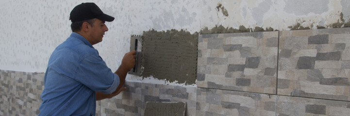 Image of a handyman tiler who with a spatula spreads the glue for laying the tiles on a house wall. Do-it-yourself work and home renovation. Horizontal banner 
