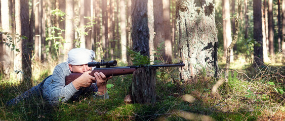 A man with an air rifle camouflages himself among the forest plants and takes aim through an...