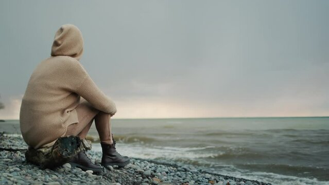 Back view: Alone woman in a warm sweater with a hood on her head sits on the ocean shore, where the dramatic sky and surf