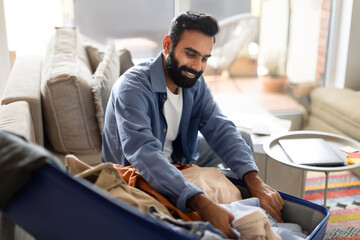 Arab bearded man near open suitcase packing clothes at home