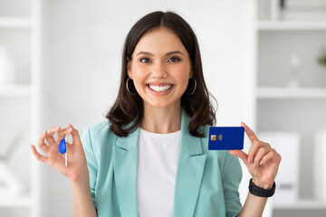 Smiling millennial caucasian lady in suit hold credit card and keys from house in office interior