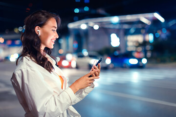 Smiling woman walking in the city at night with earphones, listening music from smartphone, side view, free space