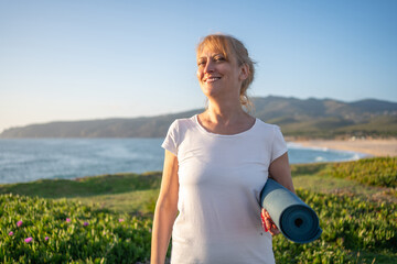 Mature woman in glasses holding yoga mat standing on beach