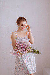 A beautiful young blonde woman in a simple white dress holds a bouquet of pale pink gypsophila flowers in her hands on a white background. Women's day concept.