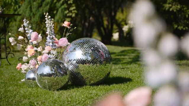 Sparkling oversized mirrored disco balls lie on green grass adorned with pastel colored flowers at themed party. Mosaic surface reflects tree on sunny holiday. Butterfly flies over decor.