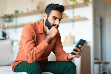 Thoughtful Indian man holding smartphone texting and thinking at home