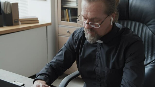 High angle shot of mature Caucasian priest wearing eyeglasses and wireless earphones, holding rosary, sitting at desk in his office typing something on laptop while having phone call