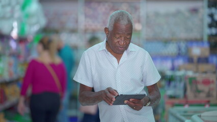 One black senior Brazilian manager of grocery store holding tablet device posing for camera inside small business. Older African American person using modern technology