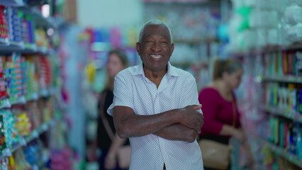 One black senior Brazilian manager of grocery store holding tablet device posing for camera inside small business. Older African American person using modern technology