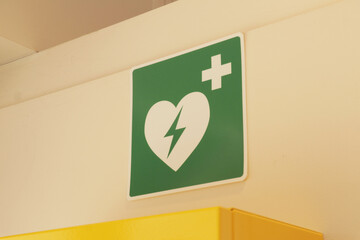 Yellow box with a defibrillator hanging on a wall with a green health care sticker sign above it. 