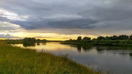 Fototapeta na wymiar The sun sinks below the horizon on a summer evening. The colorful cloudy sky and trees are reflected in the river water. The high bank of the river is overgrown with grass. A forest grows