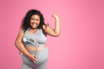 Positive black obese woman in sportswear holding scales and showing biceps, standing on pink...