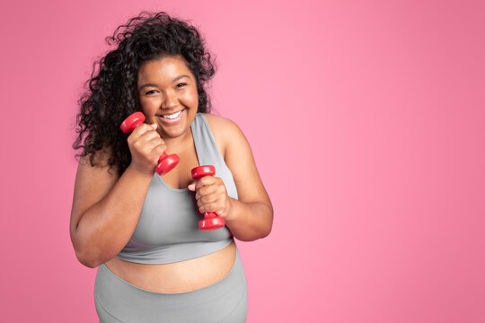 Excited plus size black woman in sportswear holding dumbbells and laughing, standing on pink background, copy space