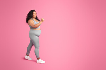 Glad black oversize woman in sportswear training, enjoying jogging and workout, pink background,...