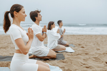 Youths practising yoga and meditation on the beach, sitting on mats with clasped hands, enjoying...