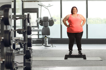 Full length portrait of a smiling corpulent woman standing on a step aerobic platform at a gym