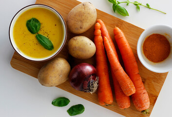 Purple bowl of potato and carrot cream soup with ingredients on wooden cutting board.