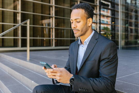 Young professional african american business man using mobile phone sitting at building office entrance.