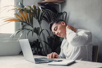 Exhausted young Caucasian male worker sit at desk massage neck suffer from strain spasm muscles. Tired unwell man overwhelmed with computer work sedentary lifestyle struggle with back pain or ache.. - obrazy, fototapety, plakaty