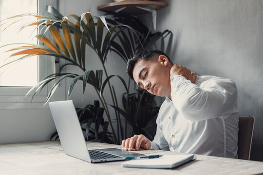 Exhausted young Caucasian male worker sit at desk massage neck suffer from strain spasm muscles. Tired unwell man overwhelmed with computer work sedentary lifestyle struggle with back pain or ache..