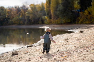 Fototapeta na wymiar a little fair-haired boy in overalls and a scarf plays on the sandy shore of the lake in autumn, a happy childhood, a natural background, a cheerful child walks in the park