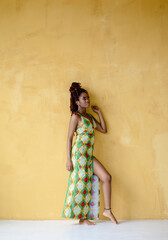 Fototapeta premium Full body photo of the young black female with a dreadlocks updo hairstyle, wearing a long ornament dress, standing barefoot, isolated on a yellow wall background with copy space.