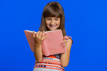 Fototapeta premium Young school girl reading funny interesting fairytale story book, leisure hobby, knowledge wisdom, education, learning, study, wow. Preteen Caucasian child kid isolated on studio blue background