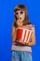 Excited young school girl in 3D glasses eating popcorn, watching interesting tv serial, sport game, film, online social media movie content. Teen female child kid on studio blue background. Vertical