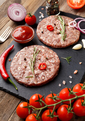 Raw beef hamburger patties on stone plate, vegetables and spices, wooden background.