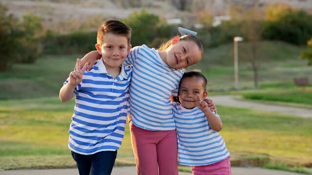 Tres ni&ntilde;os hermanos amigos primos latinos sonriendo felices en el verano al atardecer en el parque al exterior un d&iacute;a soleado abrazados juntos disfrutando de un hermoso d&iacute;a vestidos con rayas azules 