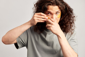 Young handsome long-haired guy applying under eye hydrogel patches on his face. Caucasian brown-haired millennial man practices skin care routine to keep healthy and youthful looking. Studio portrait.