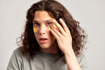 Young handsome long-haired guy applying under eye hydrogel patches on his face. Caucasian brown-haired millennial man practices skin care routine to keep healthy and youthful looking. Studio portrait.