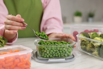 Woman taking green peas with spoon from glass container at white marble table in kitchen, closeup. Food storage