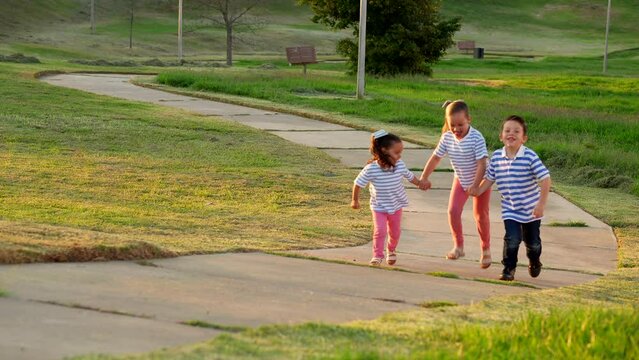 Tres ni&ntilde;os amigos hermanos latinos felices jugando y disfrutando corriendo juntos tomados de la mano cari&ntilde;osos subiendo una colina un camino sendero al aire libre en un hermoso atardecer en el parque