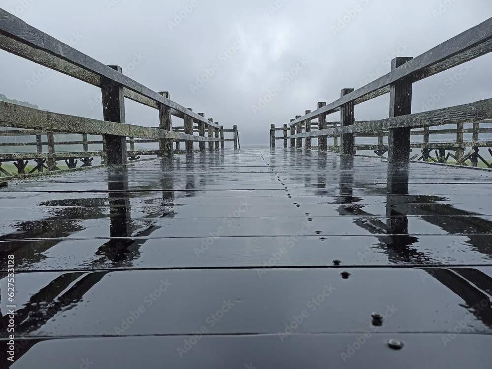 Wall mural blurry background of old and wet wooden empty bridge with gray sky over the lake on an rainy day wit