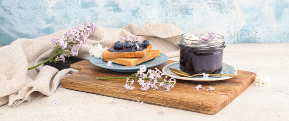 Board with jar of sweet blueberry jam, delicious toasts and flowers on table