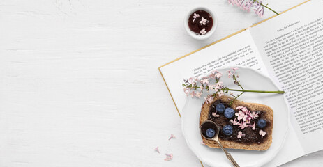 Book, plate with sweet blueberry jam toast and flowers on white wooden background with space for...