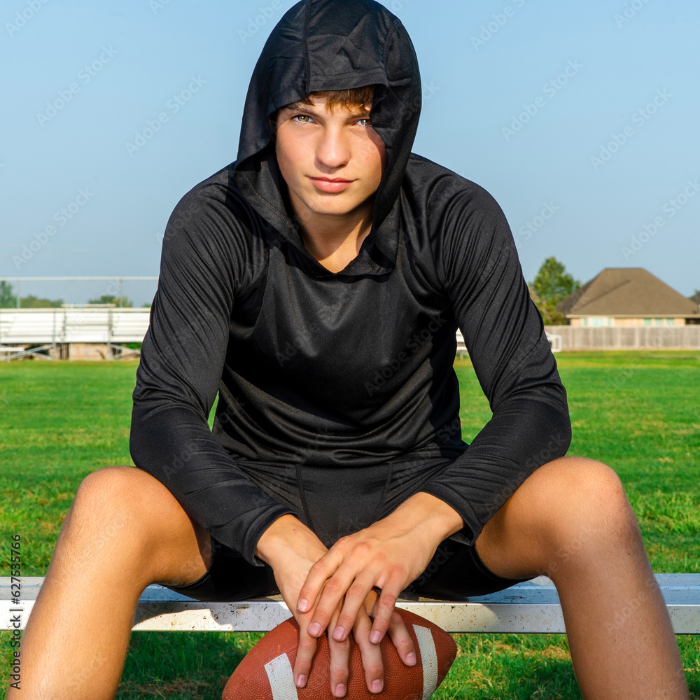 Sticker Male teenager football player in black hooded shirt sitting on bench holding a football