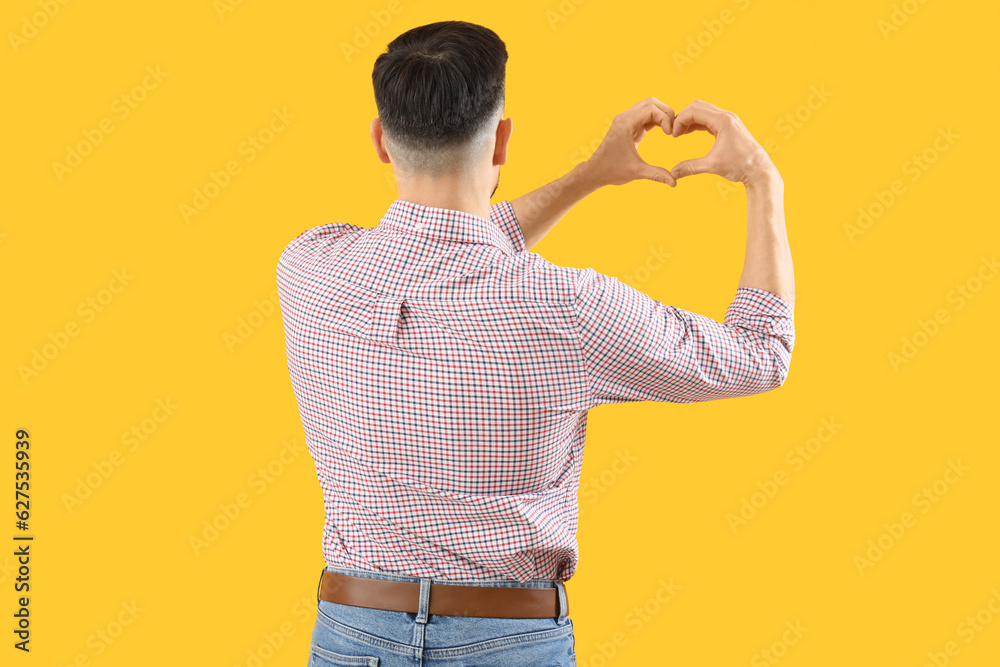 Poster handsome man making heart with his hands on yellow background, back view