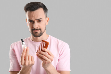 Worried young man with comb and serum for hair growth on grey background