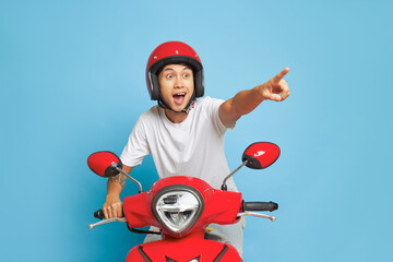 Excited young man in white t-shirt and red helmet riding red motorbike on blue background, pointing...