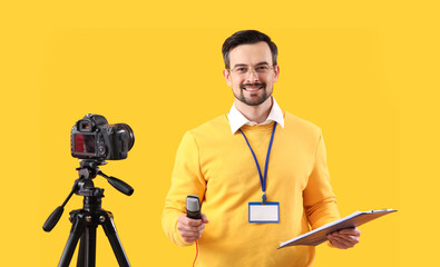 Male journalist with microphone and clipboard recording video on yellow background