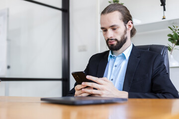 Fototapeta na wymiar Young caucasian handsome businessman with beard using smartphone sitting office table. Personal mobile communication. Man send message or search browsing information. Ordering work via online system.