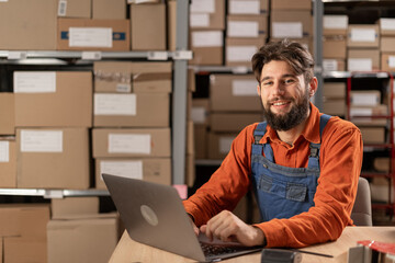Warehouse worker with laptop entering inventory data to database, smiling and looking at camera