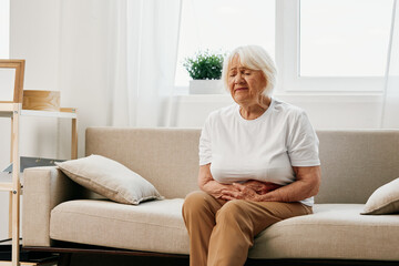 Elderly woman severe abdominal pain sitting on the sofa, health problems in old age, poor quality...