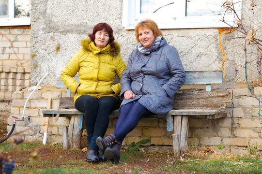 2 mature sisters sit on a bench near a village house and have a nice conversation on an autumn cold day