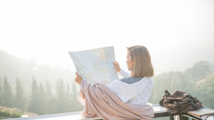 Girl tourist in mountain read the map and looking mountain view at countryside homestay in the morning sunrise with copy space. trip and relaxing concept. Woman turning back winter coat and blanket