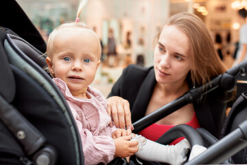 Close-up portrait of a small child in a carriage, blonde baby girl in pink clothes. Mom with a baby in a stroller walks around the mall, sat down next to the pram and talks to her daughter.