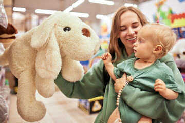 Happy young mother with her cute daughter walking around toy store in shopping mall. Mom and little...