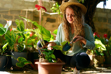 Young woman gardener in straw hat holding hand shovel taking care of potted plants. Junior caucasian female smiling sitting in her little garden planting flowers in pots. Gardening and farming concept